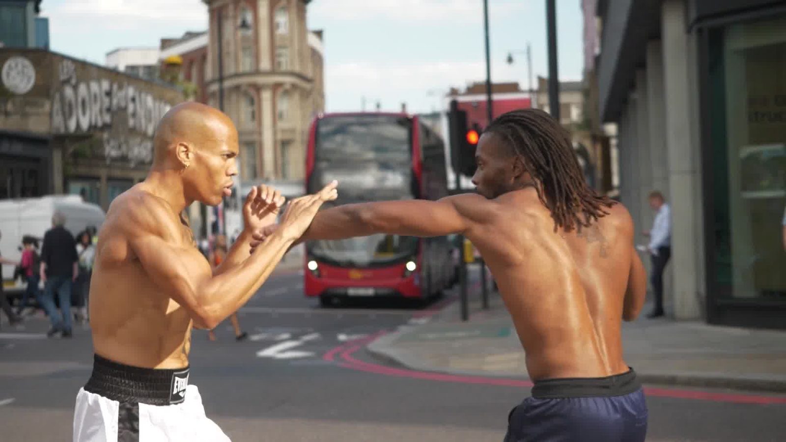 Two shirtless men spar in the middle of a London street, a red double-decker bus behind them