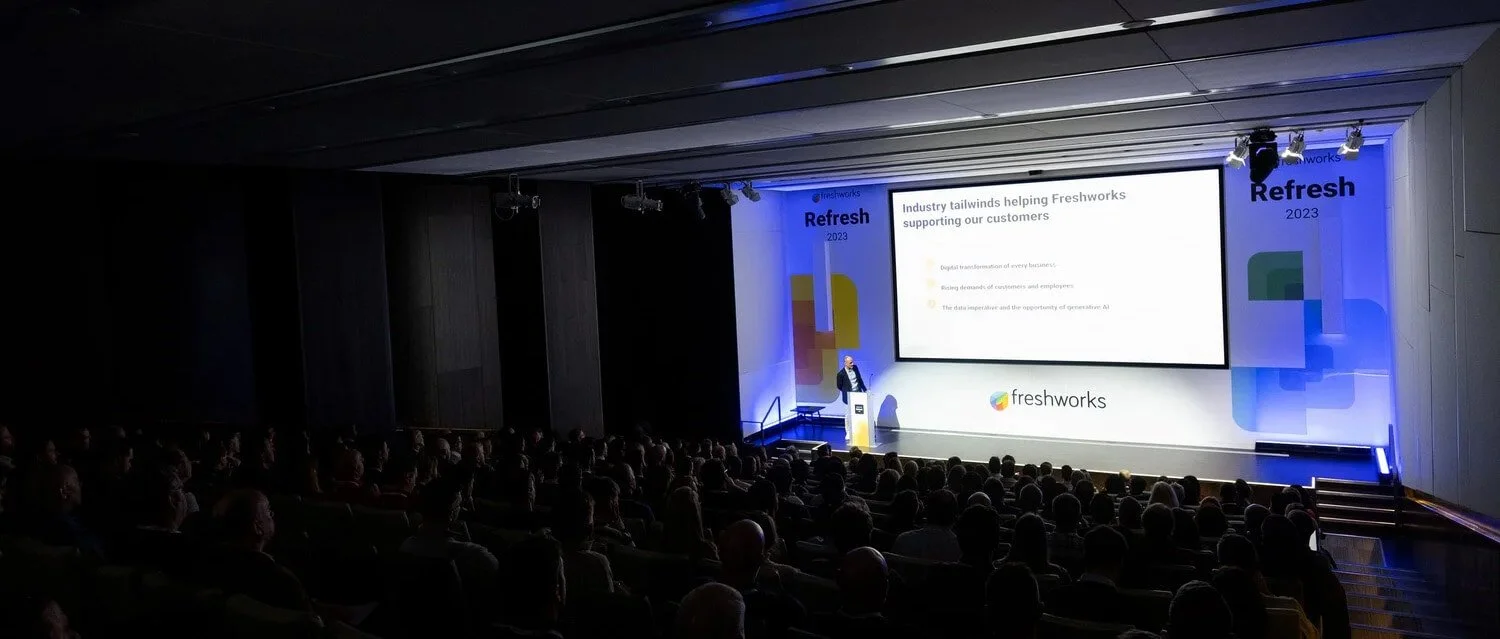 Speaker presenting on stage at the Freshworks Refresh conference in London, addressing a large seated audience with branding and slides visible