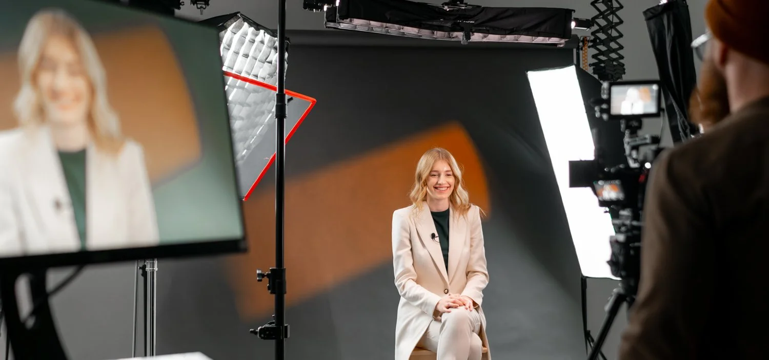 Woman in casual attire seated in a professional video recording studio, surrounded by a camera and lighting equipment.