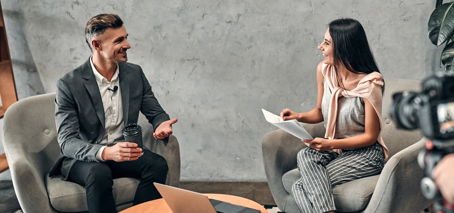 Interviewer talking with a smiling interviewee in armchairs, with a camera filming