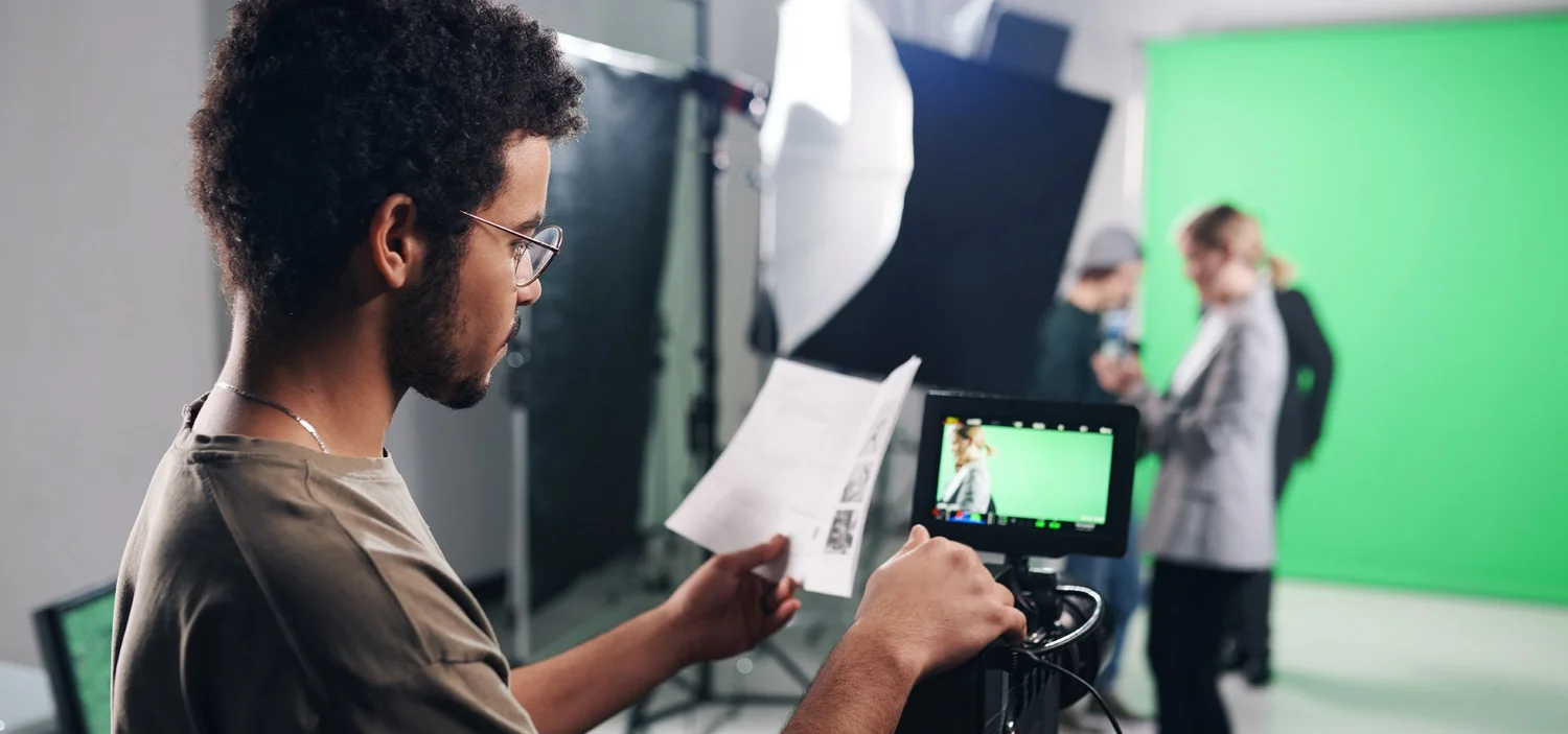 Professional video operator stood behind camera equipment, reviewing a script in a studio with a green screen background, preparing for a video shoot.